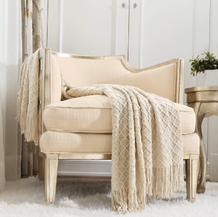 Un fauteuil élégant avec un coussin beige et un plaid en mailles sur le côté, placé près d'une table dorée dans une pièce lumineuse.